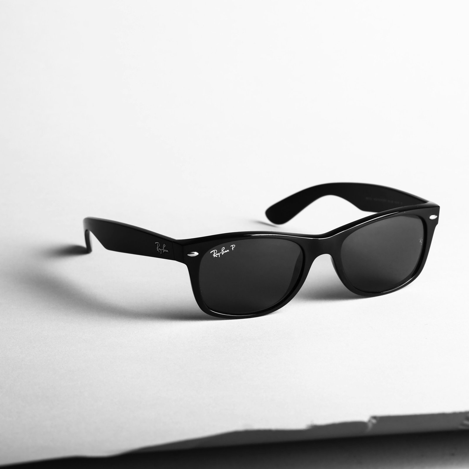 Black Ray-Ban Wayfarer sunglasses with dark lenses placed on a light surface, photographed in black and white.

